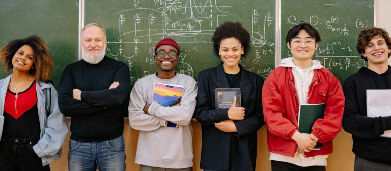 Je ziet 6 verschillende mensen op een rij staan. Van link naar rechts: een vrouw met donker krullend haar, een oudere man met wit/grijs haar, een snor en een baard, een donkere jongen met een rode muts en boeken in zijn armen, een vrouw met zwart krullend haar en boeken in haar armen, een Aziatische jongen met zwart haar en een boek in zijn handen en een jongen met donkerblond krullend haar en een notitieboek in zijn armen. Ze staan allemaal voor een groen schoolbord waar met wit krijt formules op geschreven zijn.
