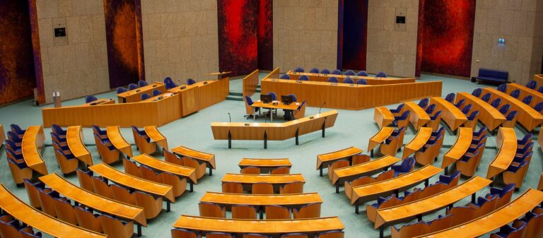 Een vergaderzaal van een parlement: een halfronde zaal met rijen houten banken en blauwe stoelen die naar een centraal spreekgestoelte zijn gericht. Achterin staan de zitplaatsen voor de voorzitter en het kabinet.