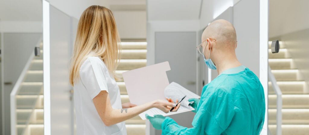 Twee zorgverleners in een medische omgeving, beide van achteren gefotografeerd. Links staat een blonde verpleegkundige in een witte blouse, die een pen en een document vasthoudt. Rechts staat een kale man in een groen operatiepak, met een mondmasker en bril. Ook hij houdt het document vast, waar ze samen naar kijken.